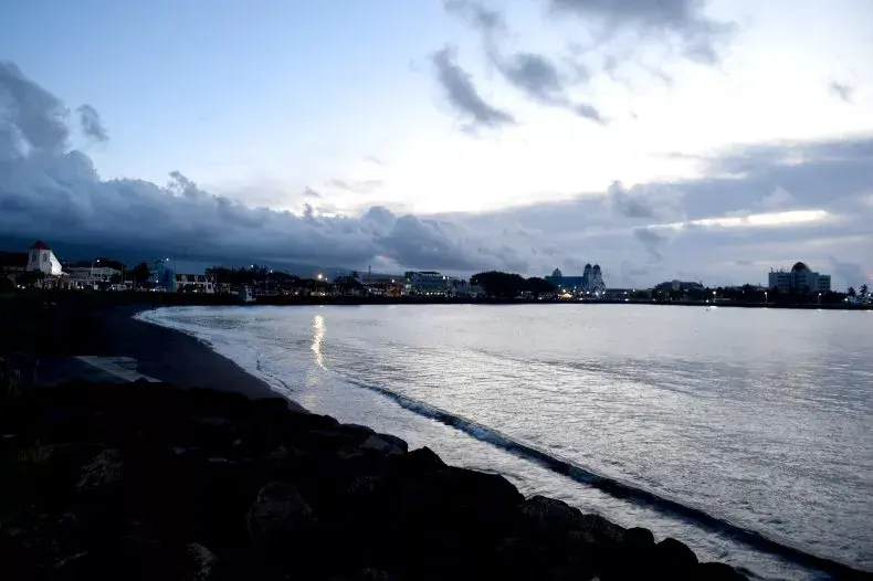 A calm coastal scene at dusk, with gentle waves and a dark shoreline in the foreground. City lights twinkle under a cloudy, moody sky in the background.